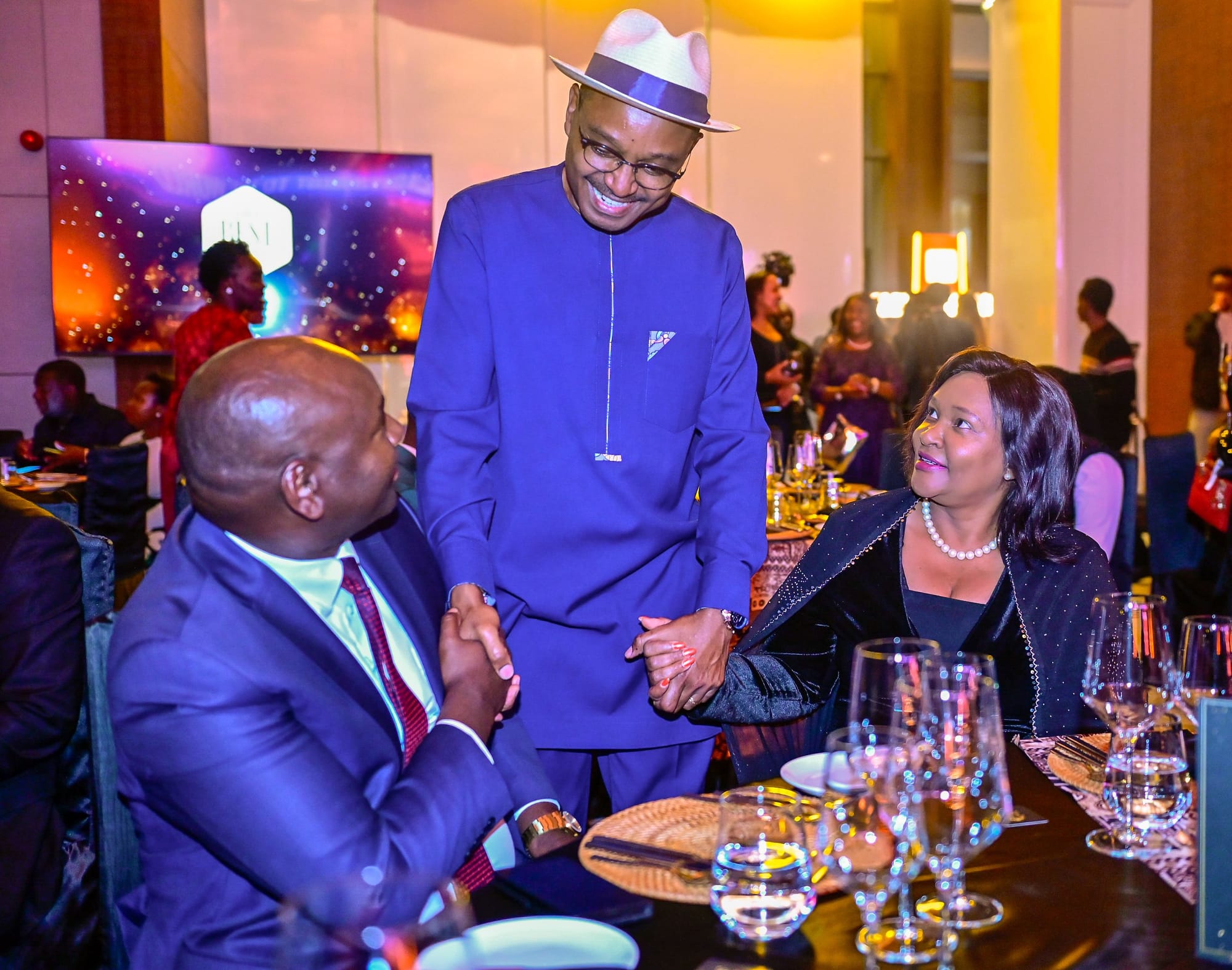 CS Rebecca Miano in a black dress smiles as a standing man in a blue tunic greets a seated guest at a dinner table.